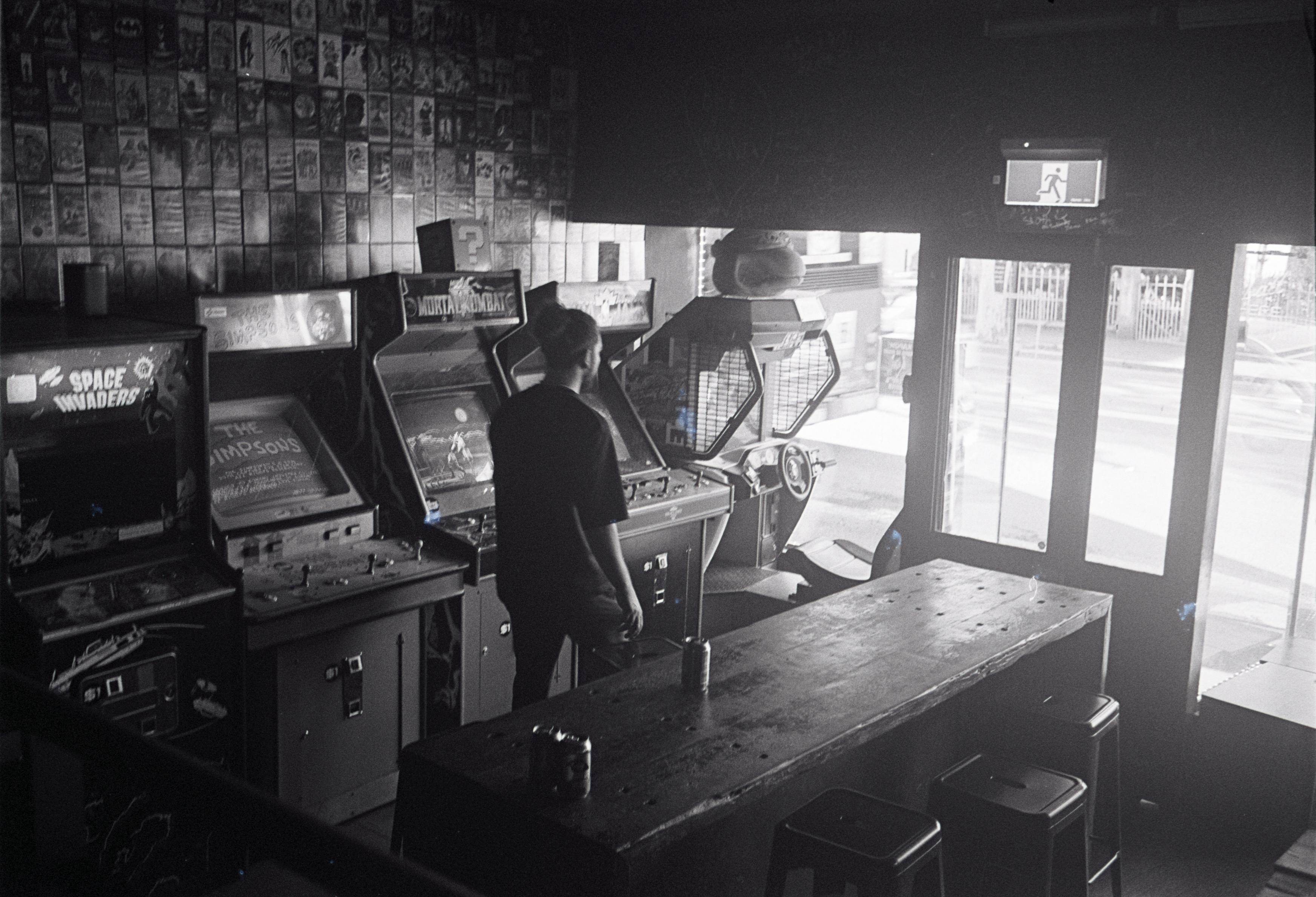A black & white photo of a video game arcade bar in Newtown, now known as Astro&rsquo;s Arcade Bar. Light spills in from outside into the dark area with the arcade cabinets.