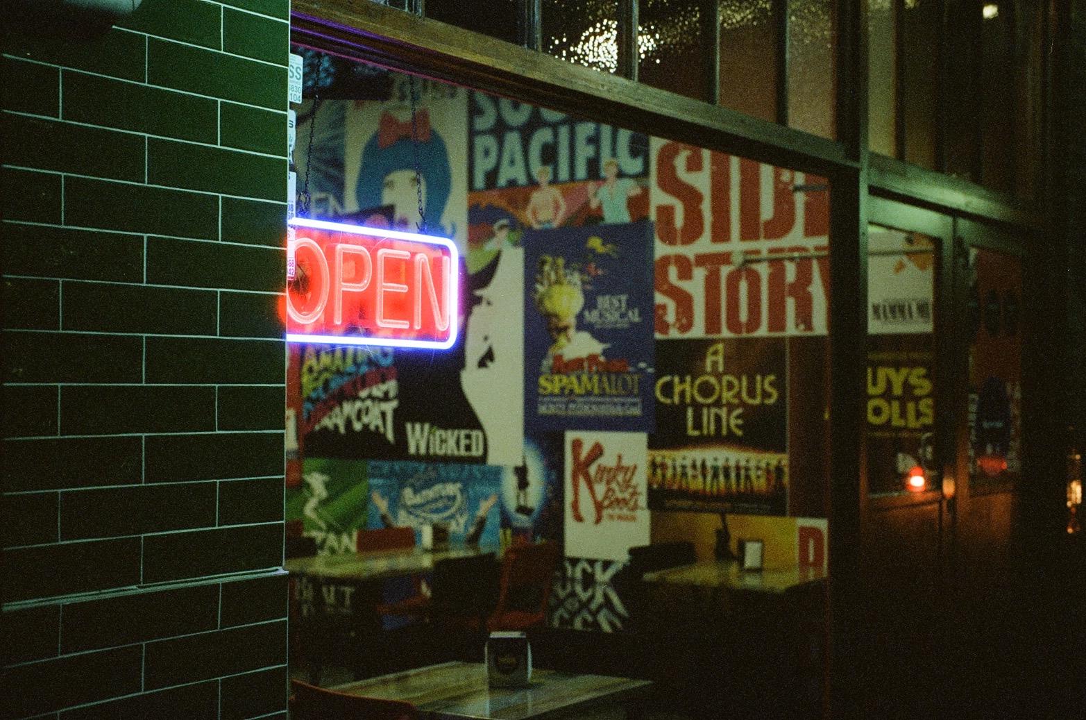 A colour photo of a burger shop at night, no visible patrons, and with a big neon OPEN sign and broadway musical posters on the far wall.