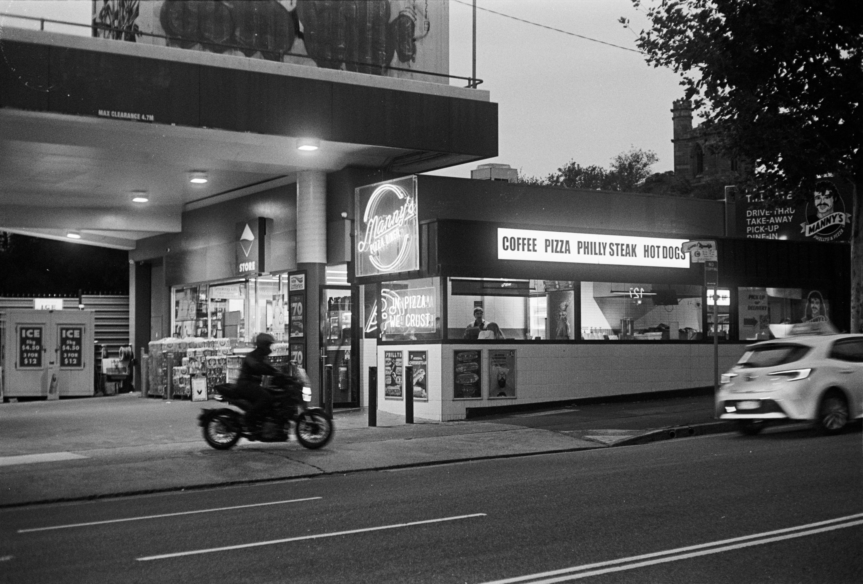 B&W photo at twilight of a pizza diner at night, some patrons inside and a bike out of focus as it blurs out of the parking lot.