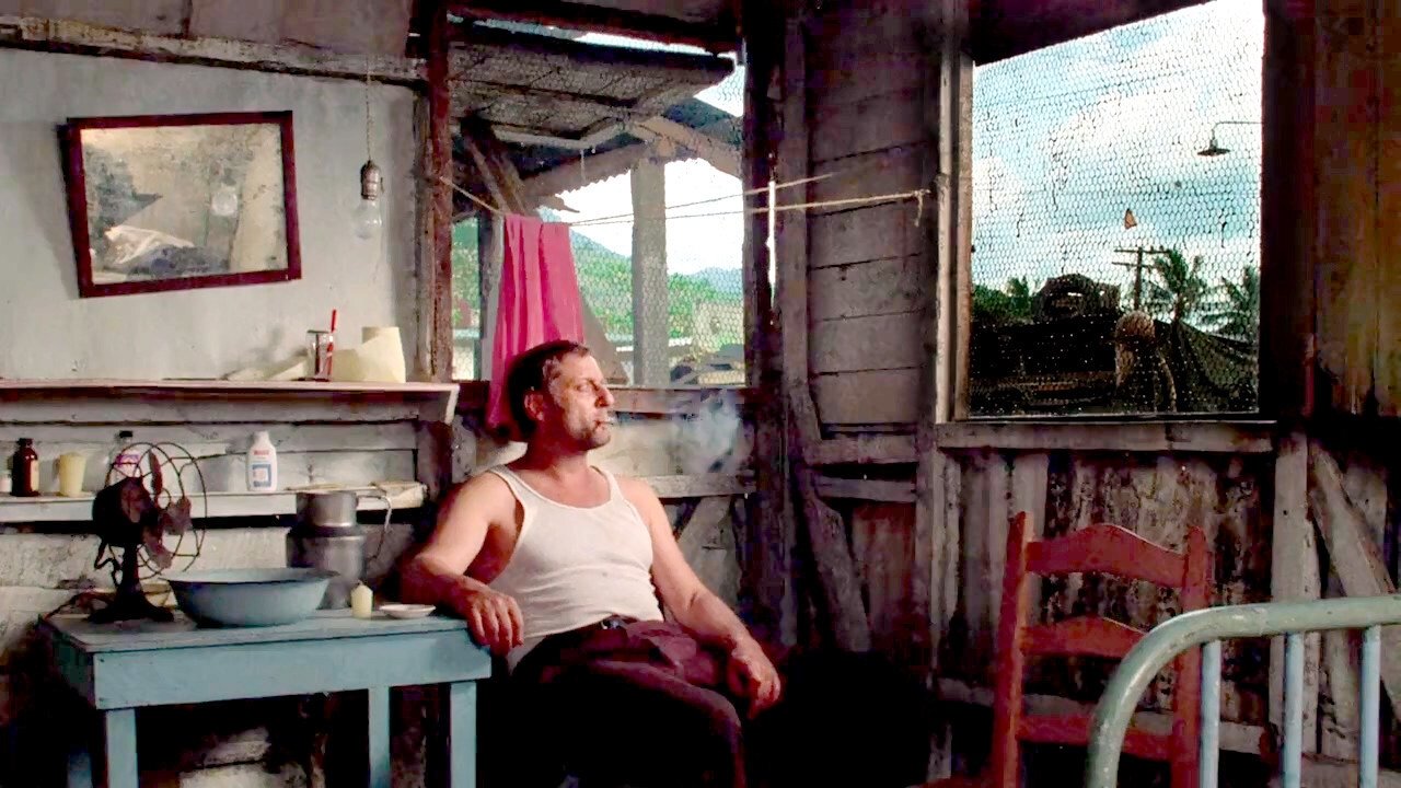 A man sits indoors in a beat-up shack in a village somewhere in South America, smoking a cigarette and bathing in his own sweat from the disgusting heat & humidity. He stares longingly out the window, no doubt thinking about the decisions that brought him to this point.